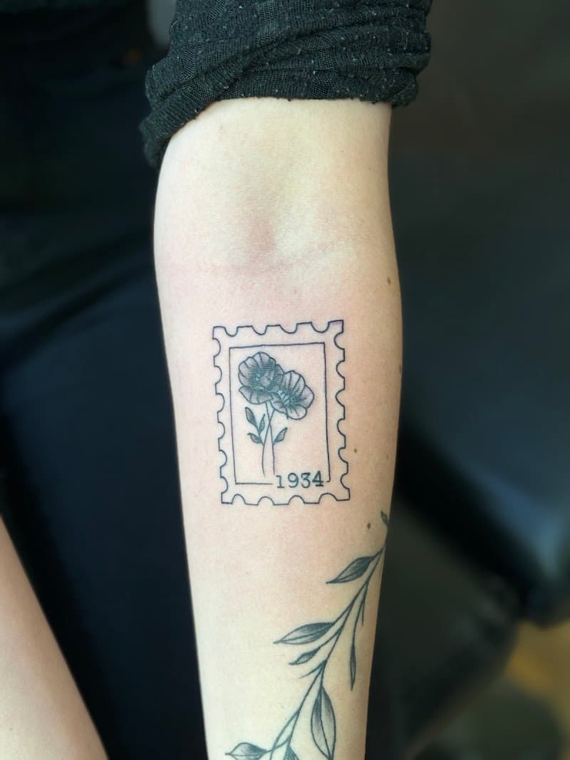 Fineline postzegel tattoo met bloemen en jaartal 1934 op onderarm.