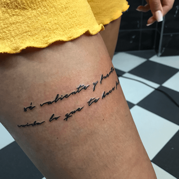 Fineline tekst tattoo op bovenbeen met Spaanse quote.