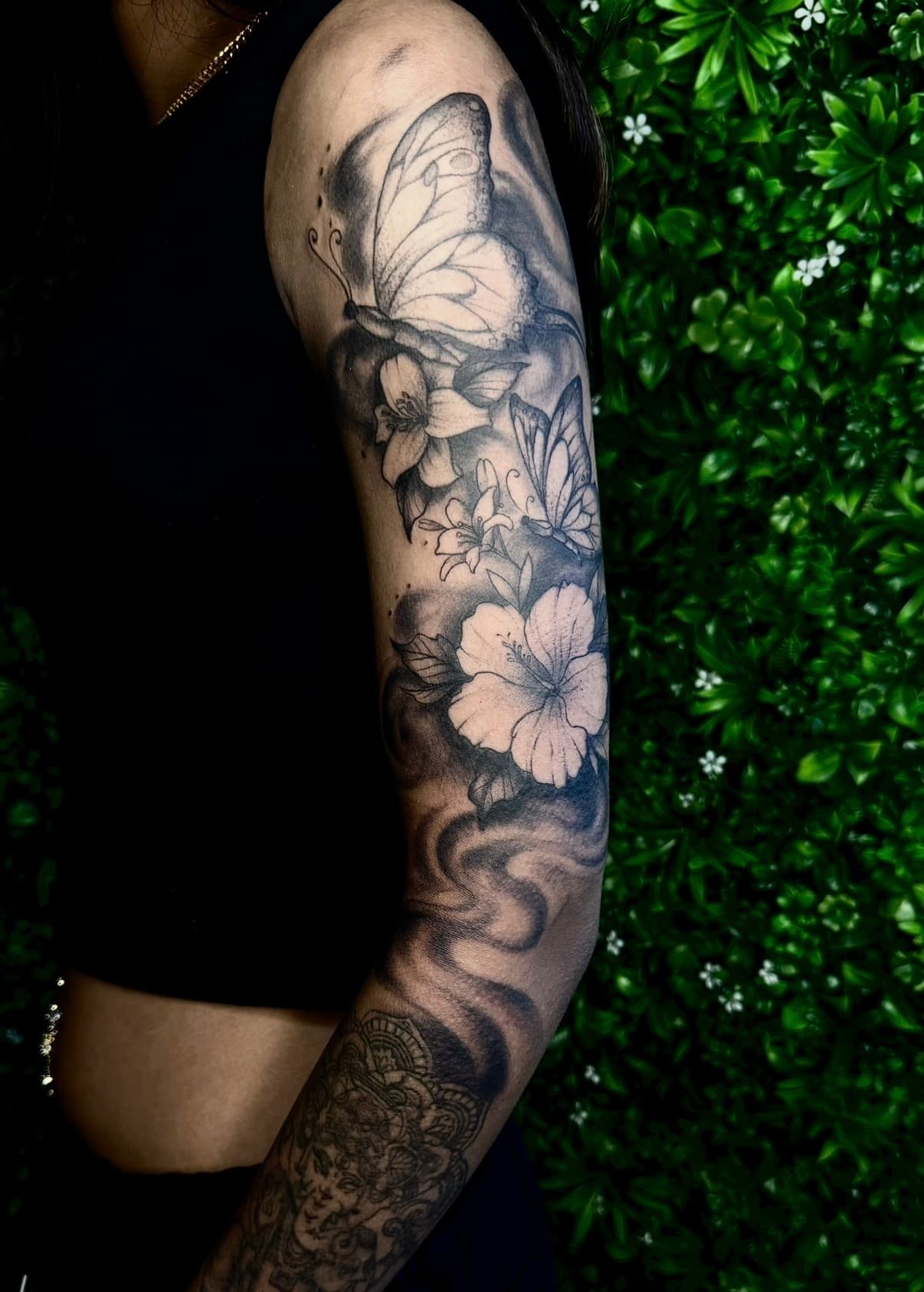 Zwart-wit tattoo van vlinders en bloemen op de bovenarm, met een mandala op de onderarm.