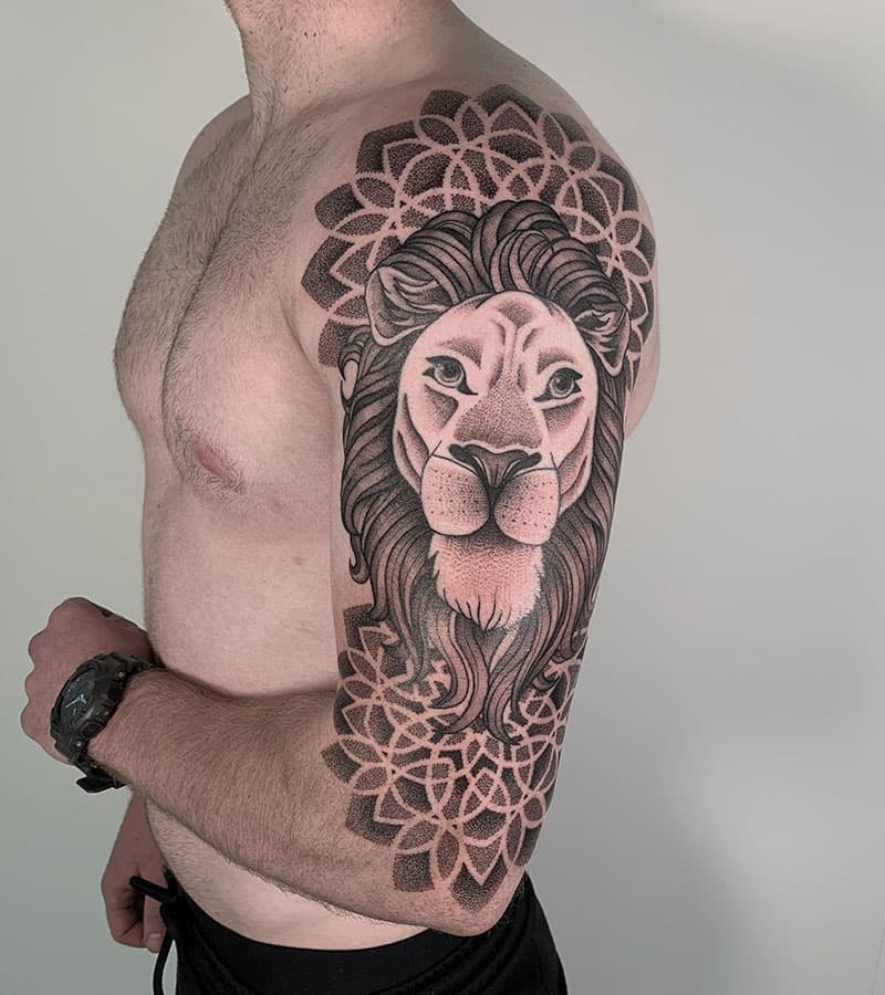 Zwart-wit tattoo van een leeuwenkop met mandala achtergrond op de bovenarm.