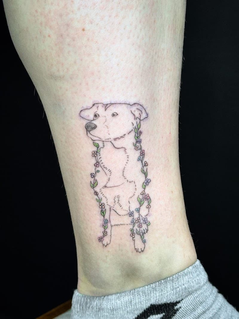 Fineline tattoo van een hond met een bloemenkrans op de enkel.
