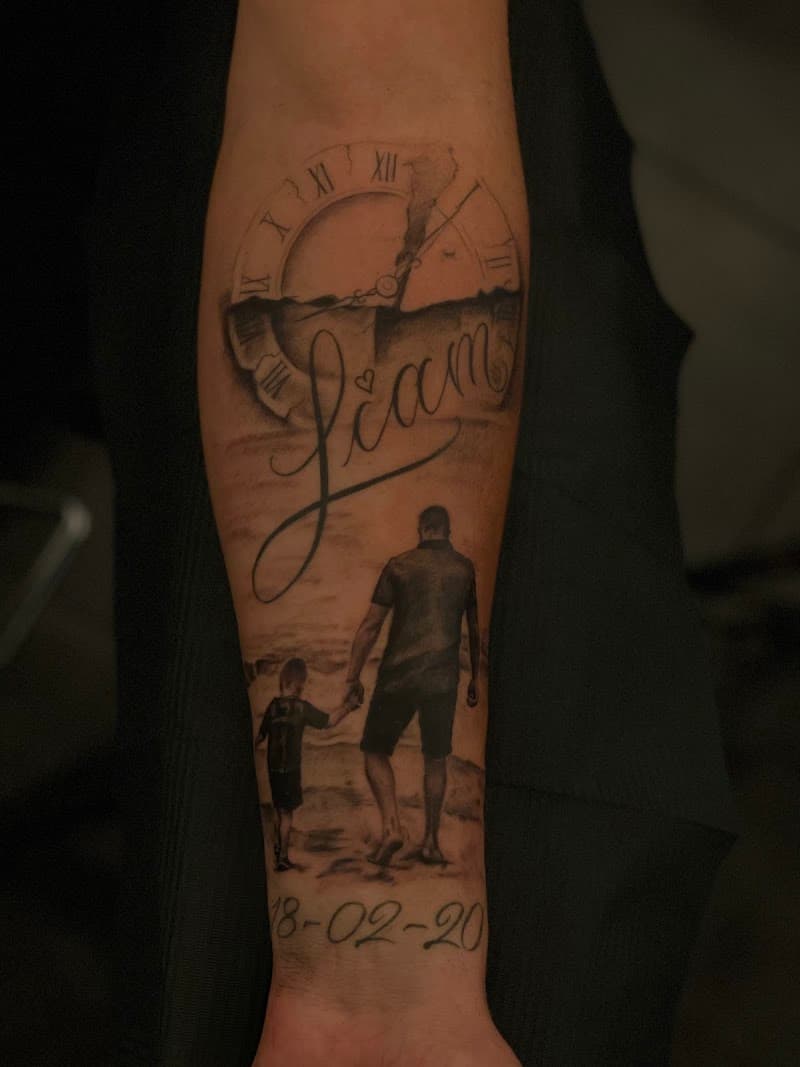Zwart-wit tattoo van vader en zoon op strand, met klok, naam 'Liam' en datum.