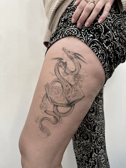 Fineline tattoo van een draak die uit een magisch boek komt op het bovenbeen.