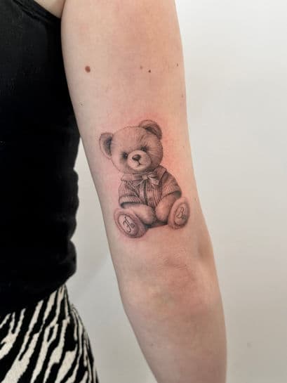 Gedetailleerde illustratieve teddybeer tattoo in zwart-wit op de bovenarm.