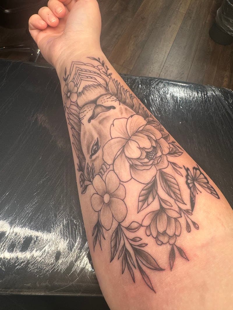 Zwart-wit tattoo op onderarm met leeuwenkop, bloemen en een vlinder.