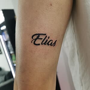 "Elias" naam tattoo in sierlijk handschrift op de bovenarm.