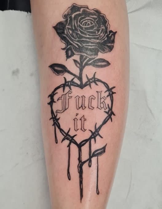 Zwart-wit tattoo van een roos in een hart van prikkeldraad met tekst 'Fuck it'.