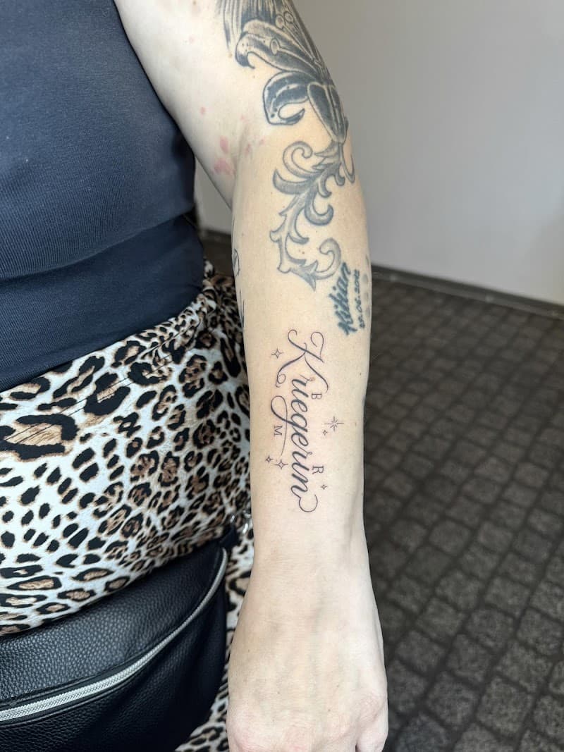 Fineline script tattoo 'Kriegerin' met sterretjes op onderarm.