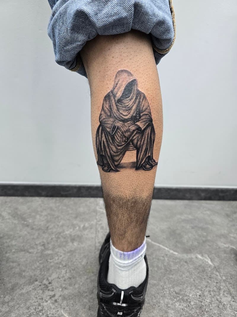 Zwart-wit tattoo van een mysterieuze zittende figuur met een kap op het onderbeen.
