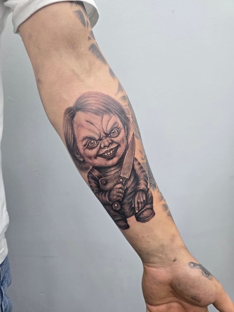 Realistische blackwork tattoo van Chucky met een mes op de onderarm.