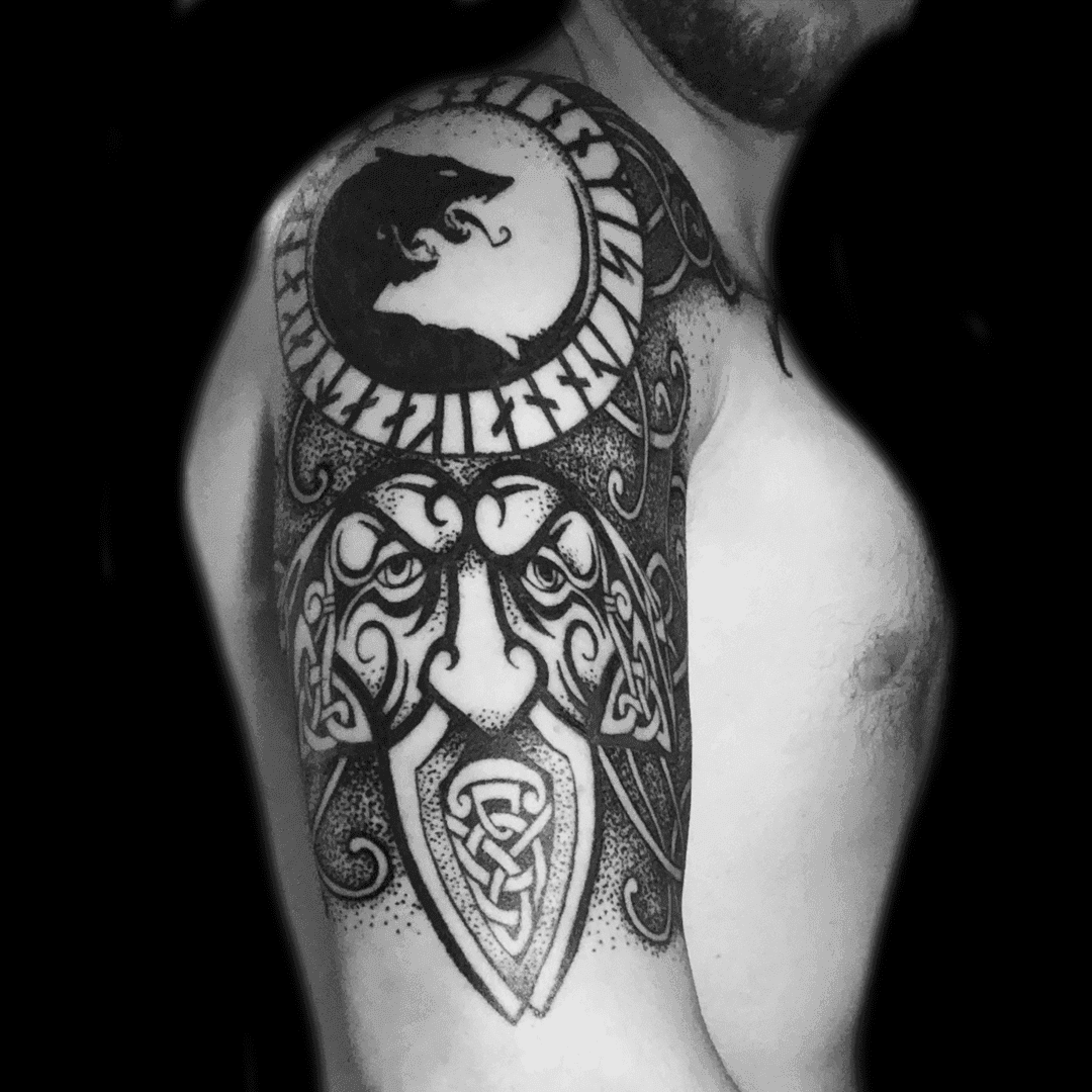 Zwart-wit tattoo van een wolf in een runencirkel en een Keltisch gezicht op de bovenarm.
