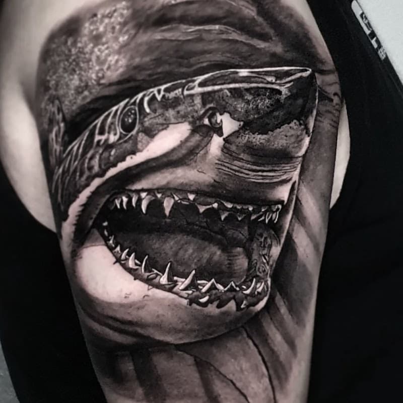 Realistische zwart-wit tattoo van een haai met open bek op de bovenarm.
