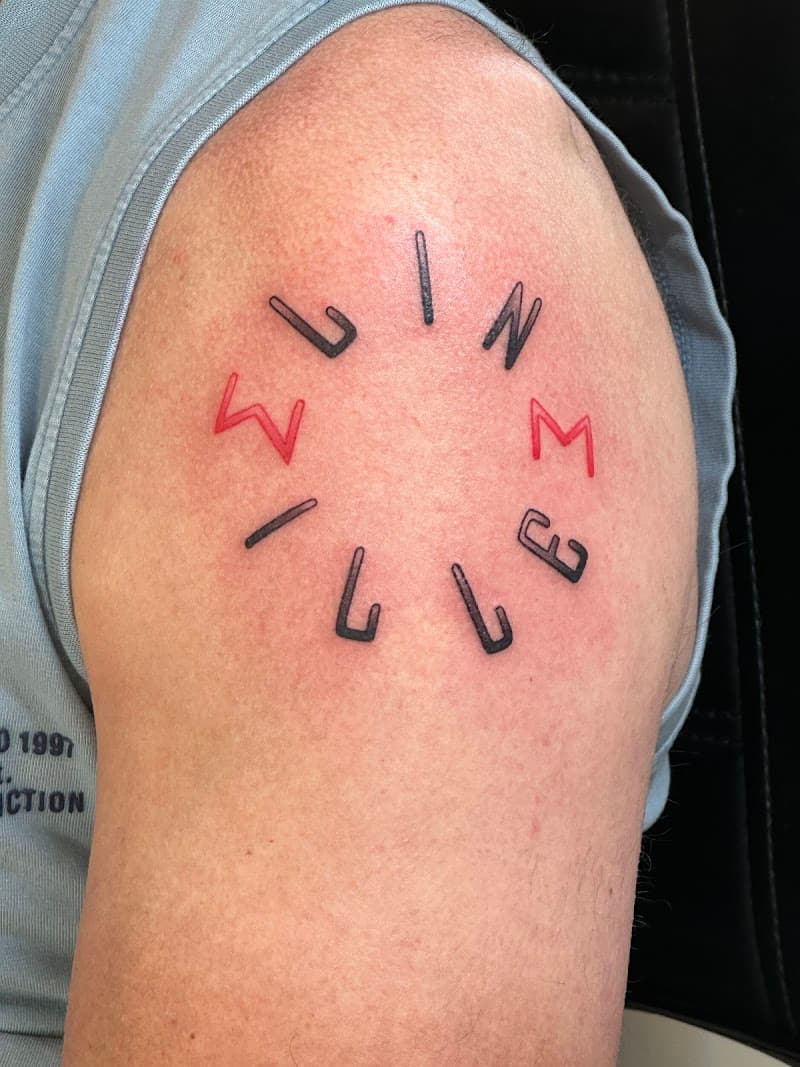 Cirkelvormige tekst tattoo met zwarte en rode letters op de schouder.