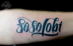Zwart-wit 'Soso lobi' tekst tattoo in sierlijk schrift op de onderarm.