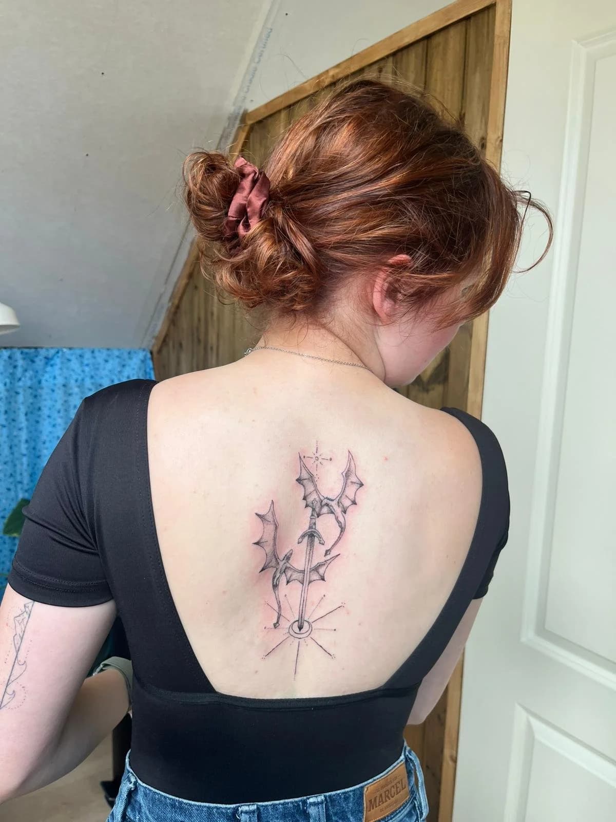 Fineline tattoo van een zwaard met drakenvleugels op de bovenrug.