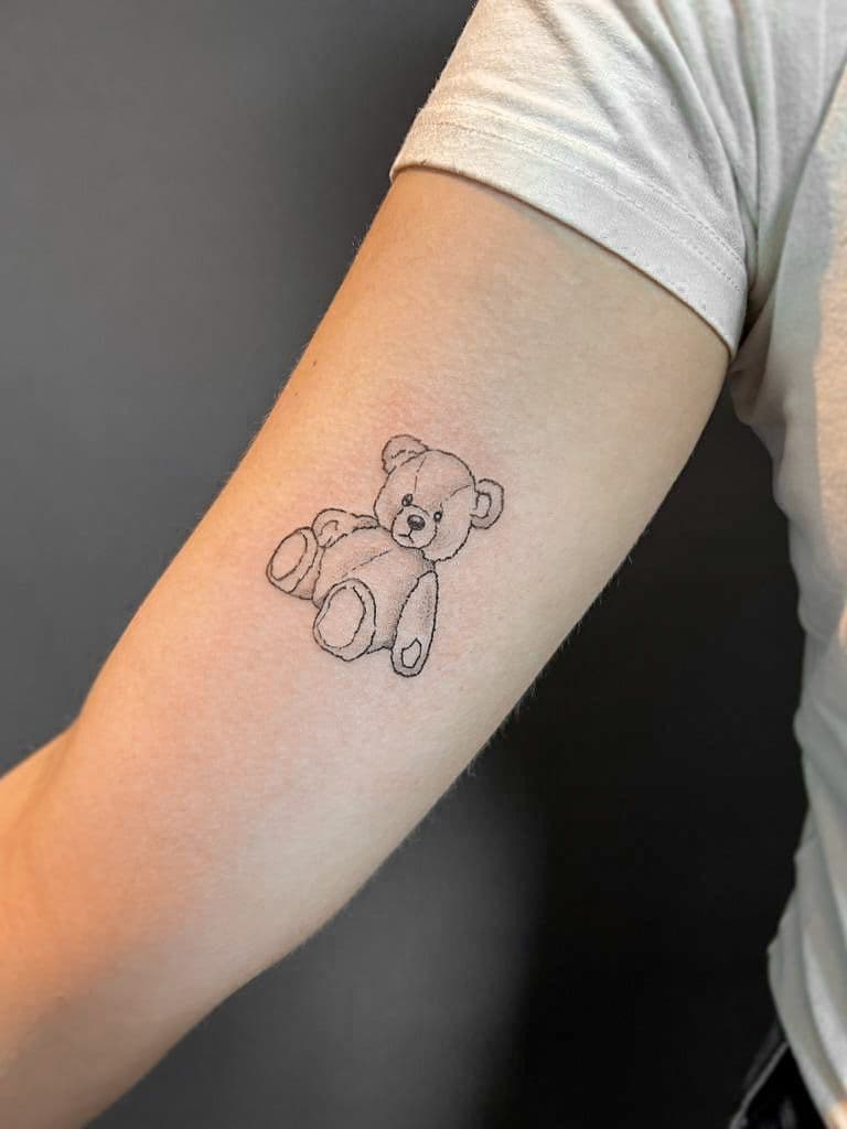 Minimalistische fineline tattoo van een teddybeer op de bovenarm.
