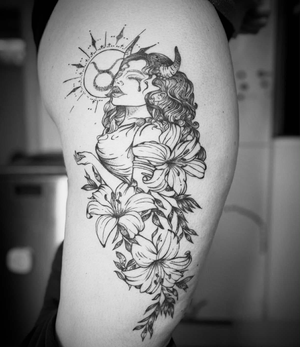 Zwart-wit tattoo van een gehoornde vrouw (Stier) met lelies op het bovenbeen.