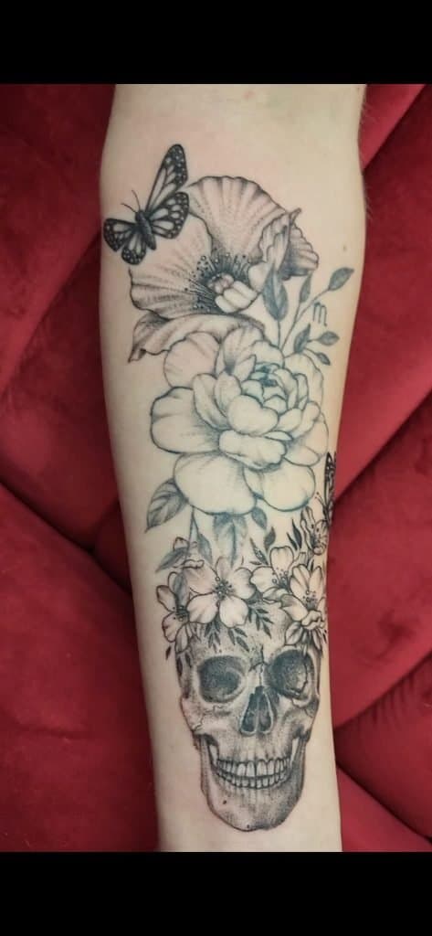 Blackwork tattoo van een schedel met bloemen en een vlinder op de onderarm.
