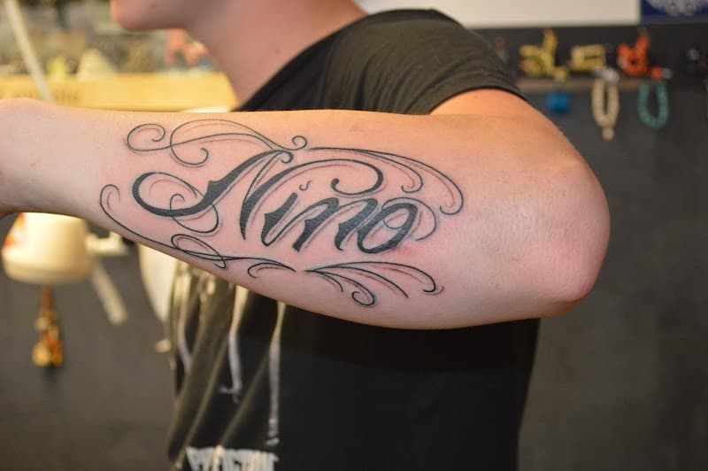 Zwart-wit lettering tattoo van de naam 'Nino' met sierlijke krullen op de onderarm.