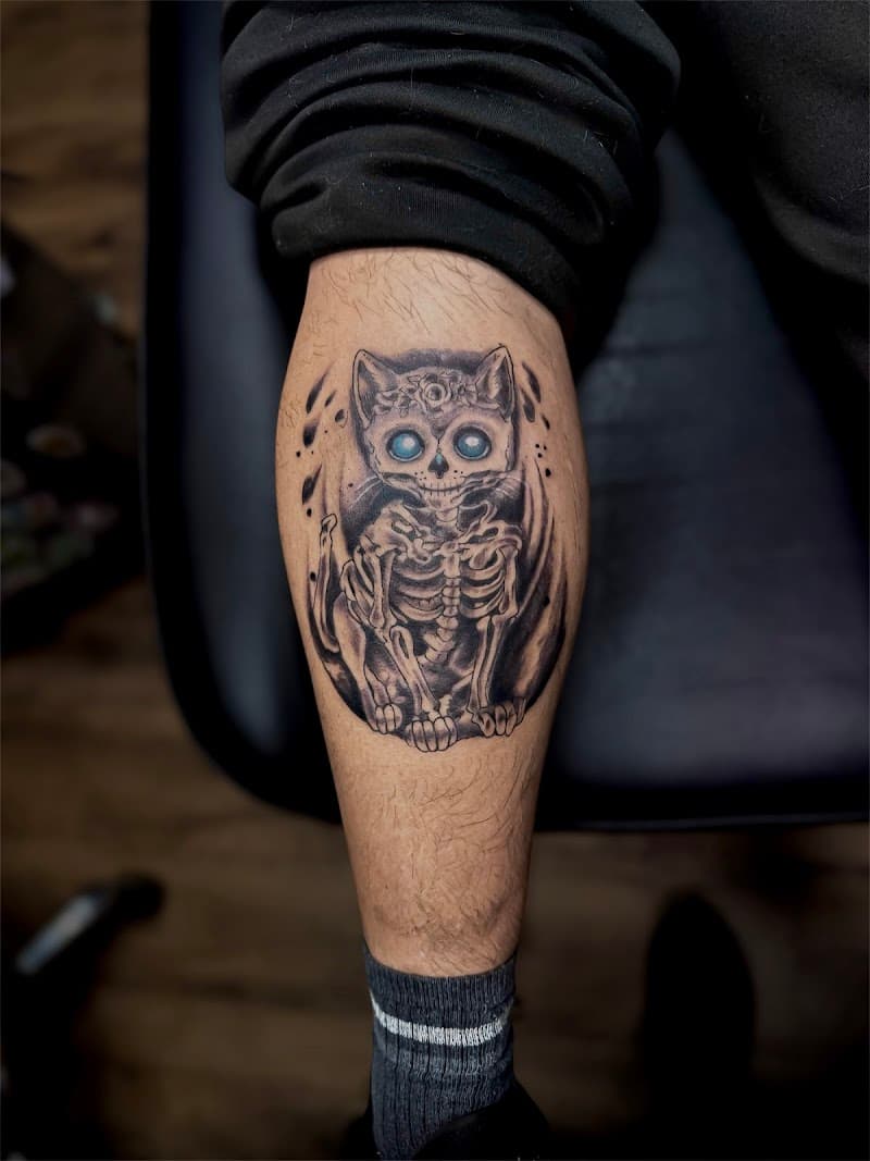 Zwart-grijze kat skelet tattoo met felblauwe ogen op onderbeen.