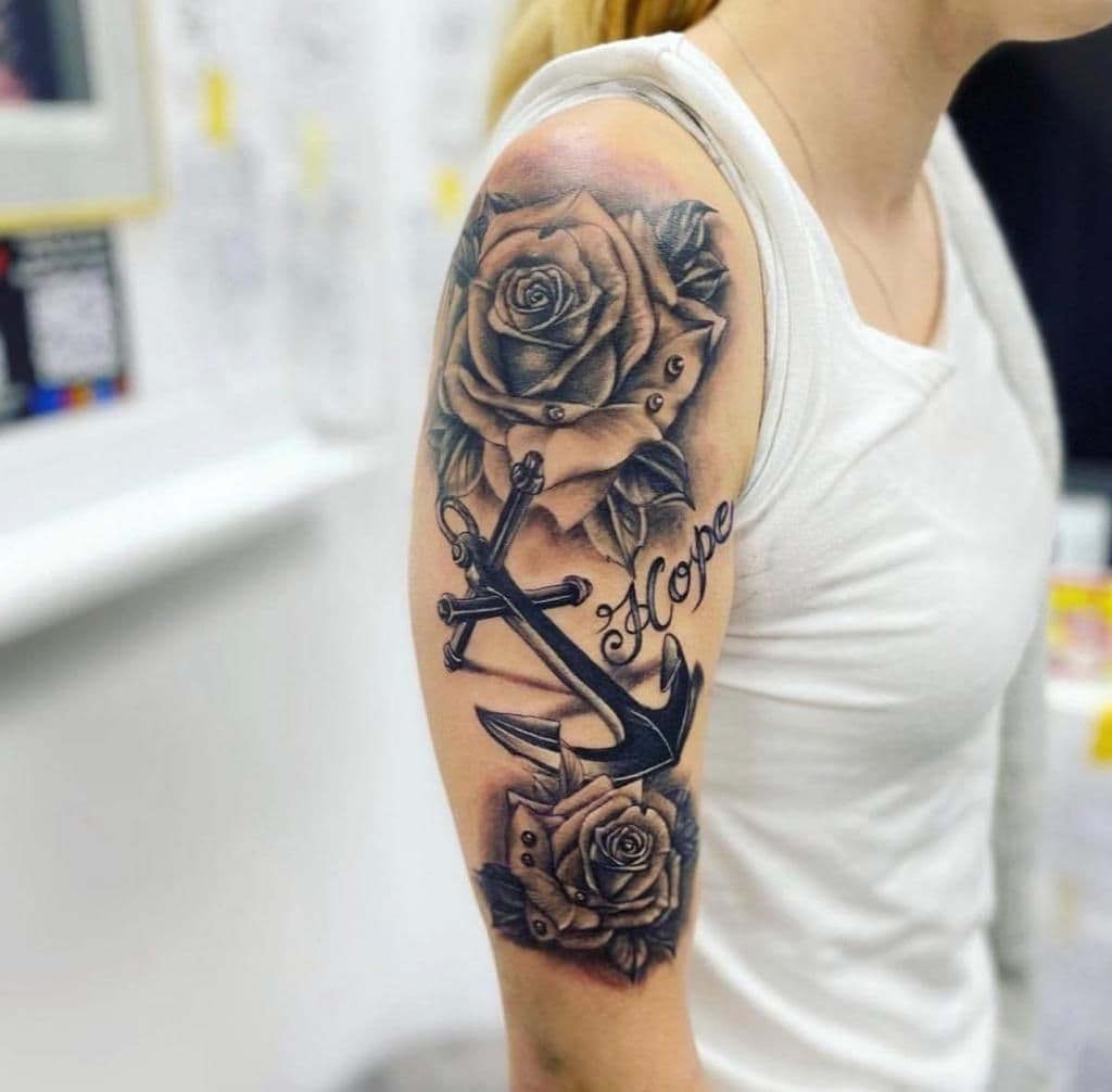 Zwart-grijze tattoo van rozen, een anker en het woord 'Hope' op de bovenarm.