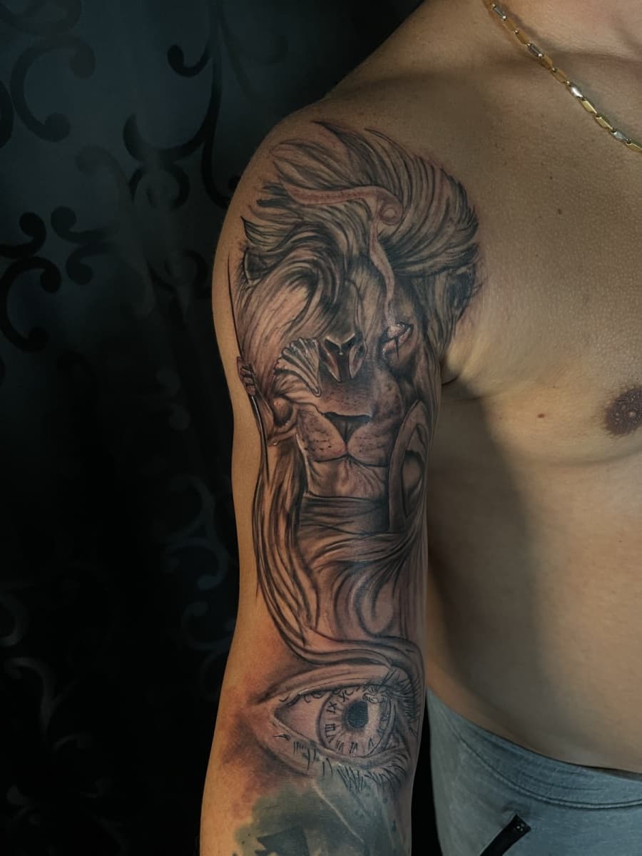 Zwart-wit tattoo van een leeuw met een oog en klok eronder op de bovenarm.