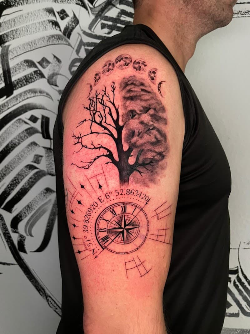 Zwart-wit tattoo van boom, maanfasen en kompas met coördinaten op bovenarm.
