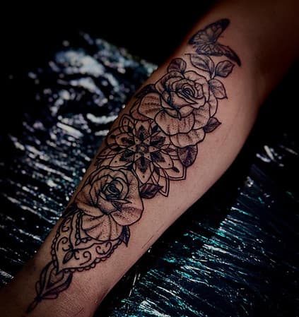 Zwart-wit tattoo van rozen, mandala en vlinder op onderarm.