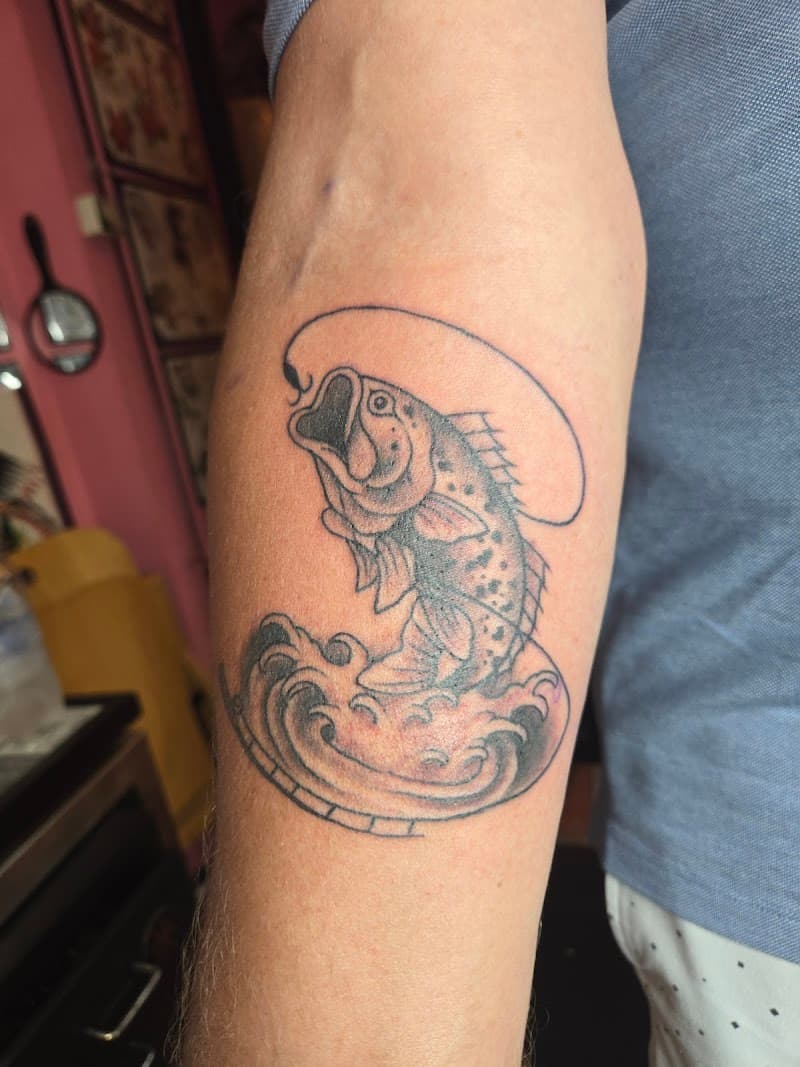 Zwart-wit tattoo van een springende vis aan een haak in golven op de onderarm.