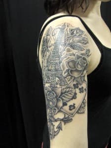 Zwart-wit tattoo van Eiffeltoren, rozen en speelkaart op bovenarm.