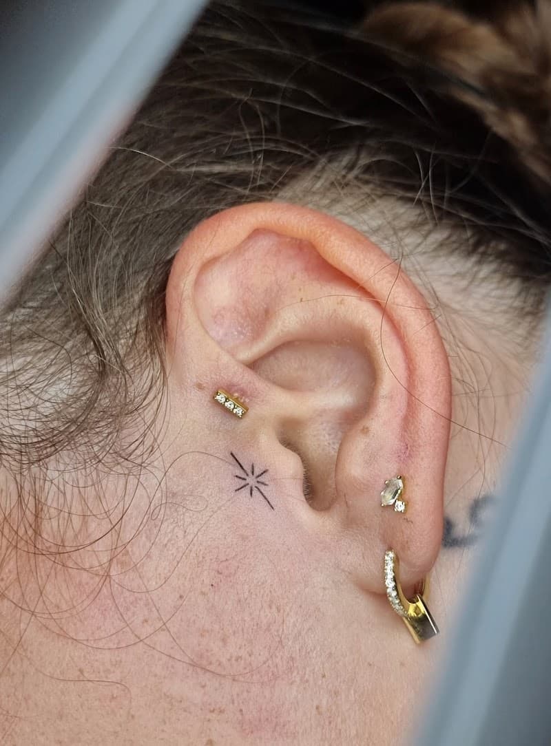 Kleine fineline ster of vonk tattoo achter het oor.
