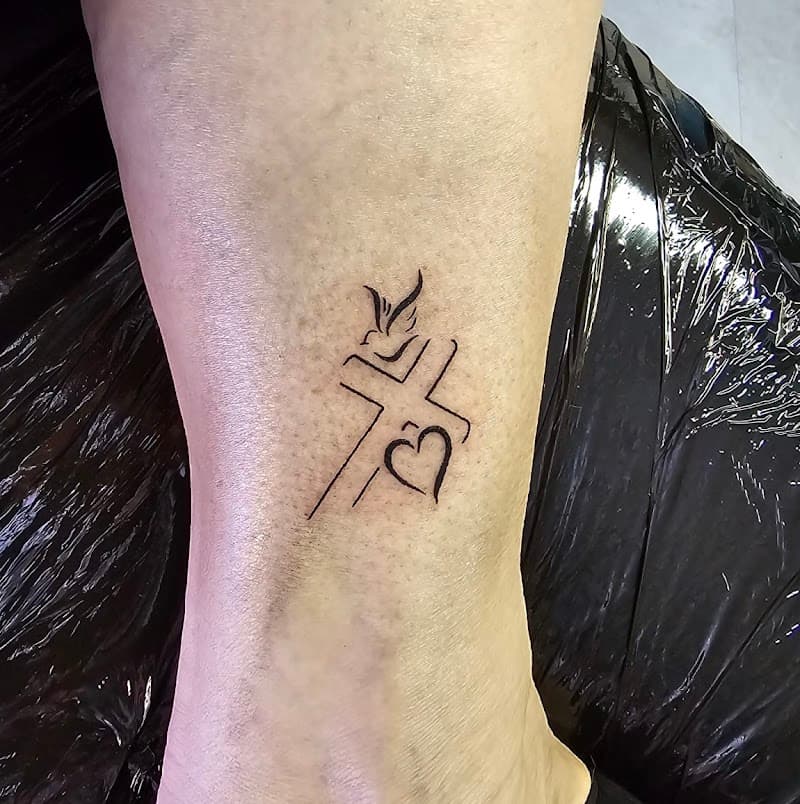 Minimalistische fineline tattoo van een kruis, hart en duif op de enkel.