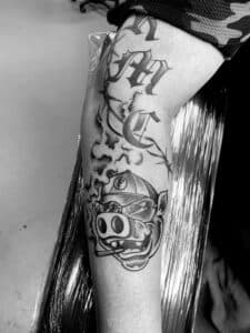 Zwart-wit tattoo van een varken met pet en sigaar, en gotische letters erboven.
