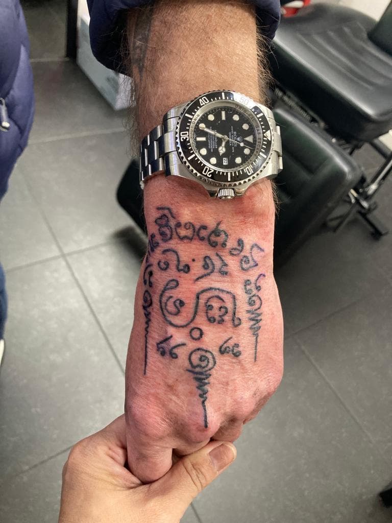 Sak Yant stijl tattoo met script en symbolen op de bovenkant van een hand.