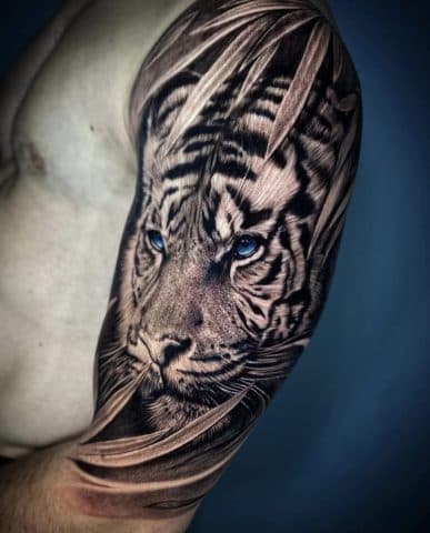 Realistische tijger met blauwe ogen tussen bladeren op bovenarm.