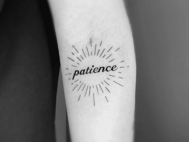 Fineline 'patience' tekst met stralenkrans op de achterkant van de bovenarm.
