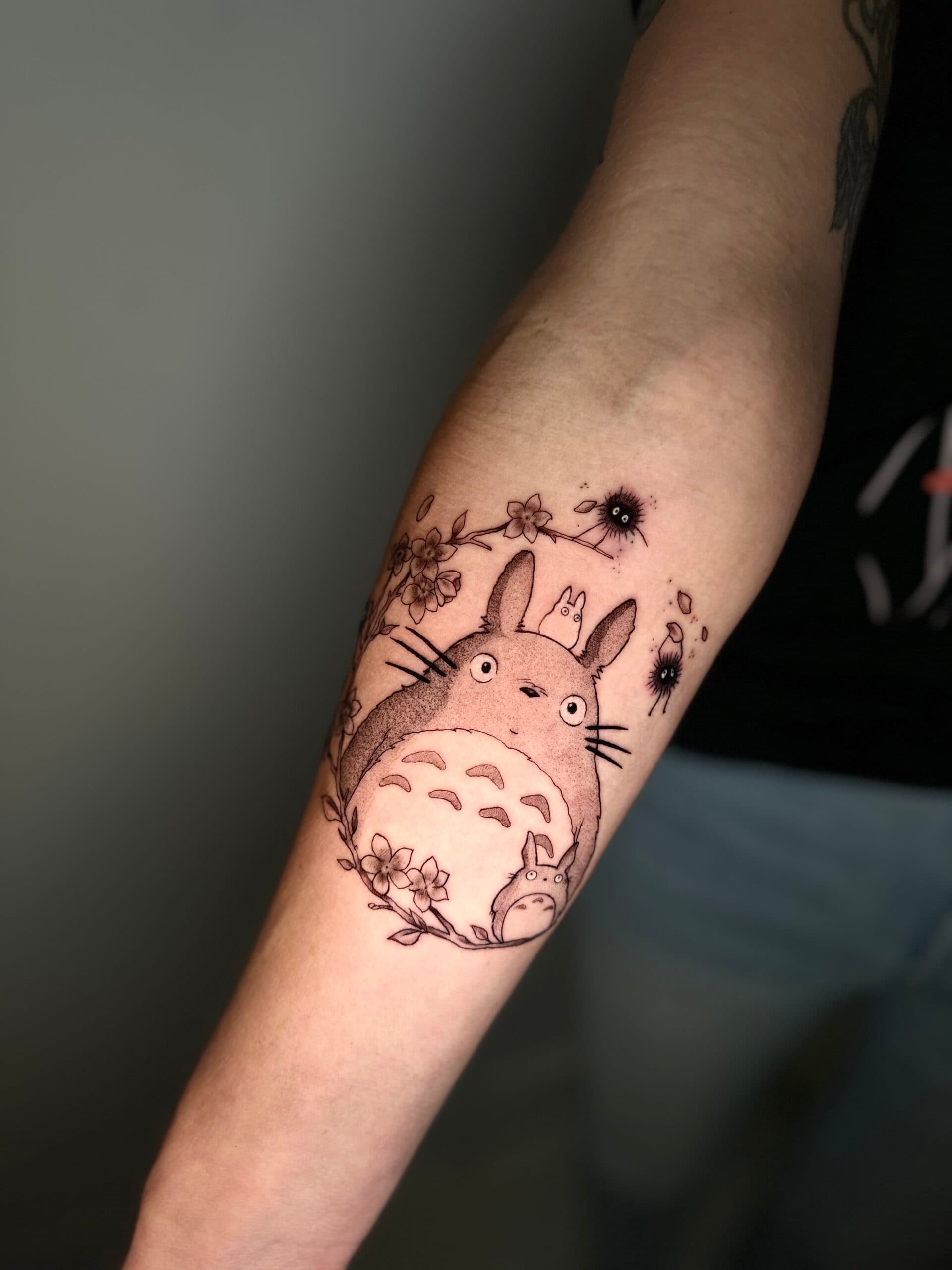 Zwart-wit Totoro tattoo met kersenbloesem en stofjes op onderarm.