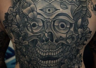 Grote zwart-wit rugtattoo van een schedel met derde oog en bloemen.