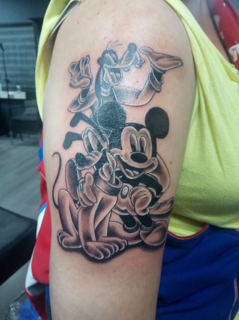 Zwart-wit tattoo van Mickey Mouse, Goofy en Pluto op de bovenarm.
