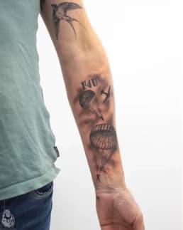 Zwart-wit tattoo van parachutisten, vliegtuig en zwaluw op arm.