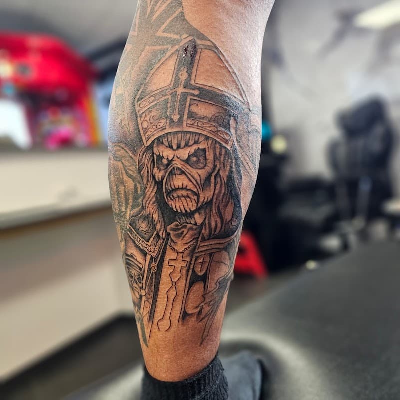 Zwart-wit tattoo van Iron Maiden's Eddie als priester op onderbeen.