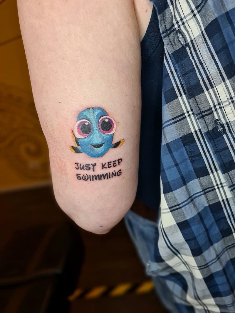 Kleine kleuren tattoo van baby Dory met tekst 'JUST KEEP SWIMMING' op bovenarm.