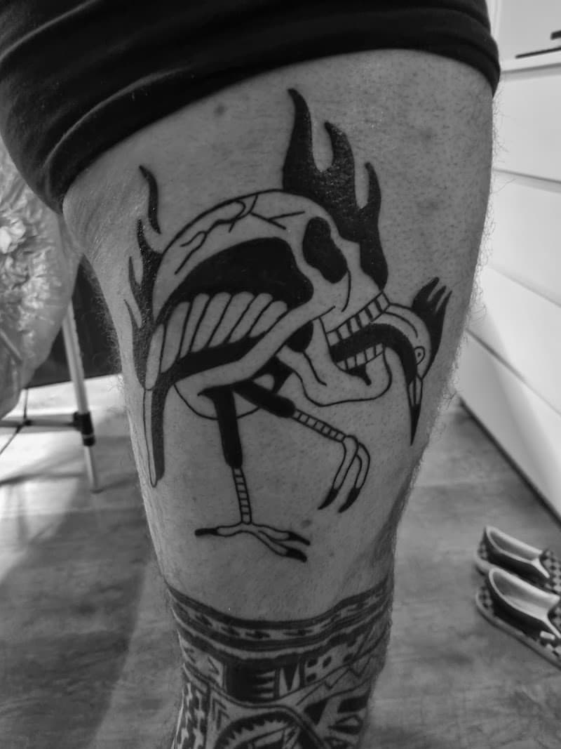 Zwart-wit tattoo van een schedel met kraanvogelpoten en vlammen op het bovenbeen.
