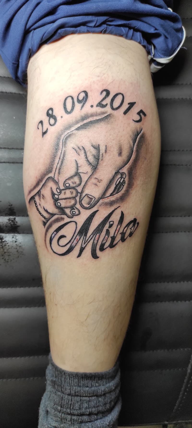 Blackwork tattoo van een volwassen hand die een babyhandje vasthoudt, met naam en datum.