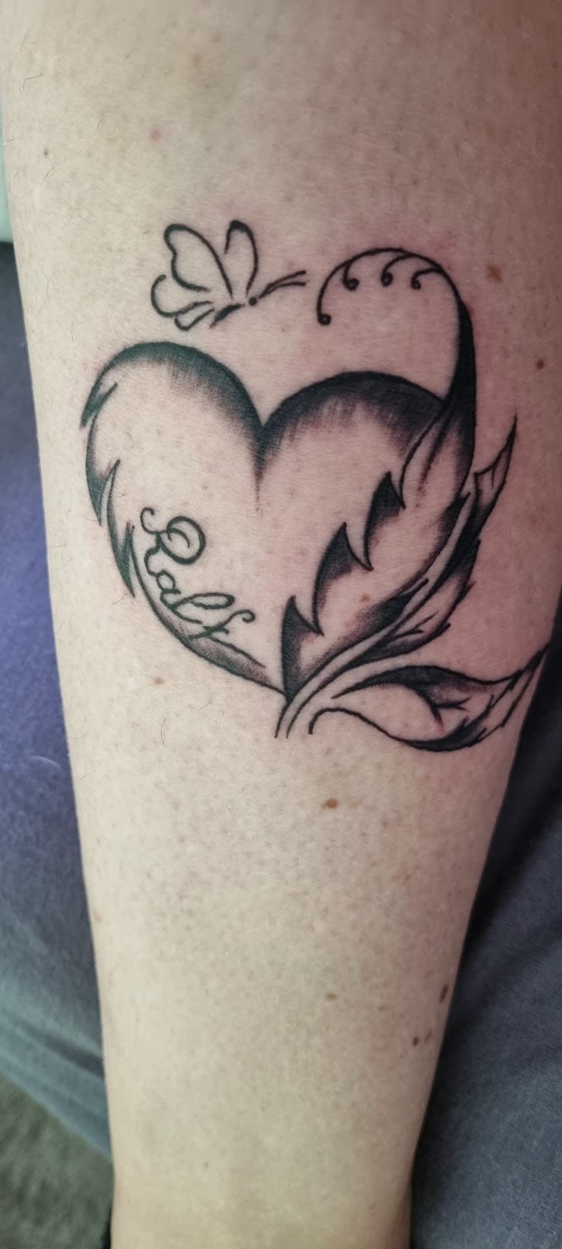 Hartvormige tattoo met veer, blad, vlinder en naam 'Ralf'.
