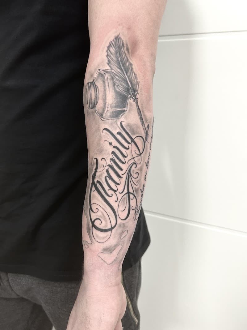 Zwart-wit tattoo van een inktpot met veer en het woord 'Family' op de onderarm.