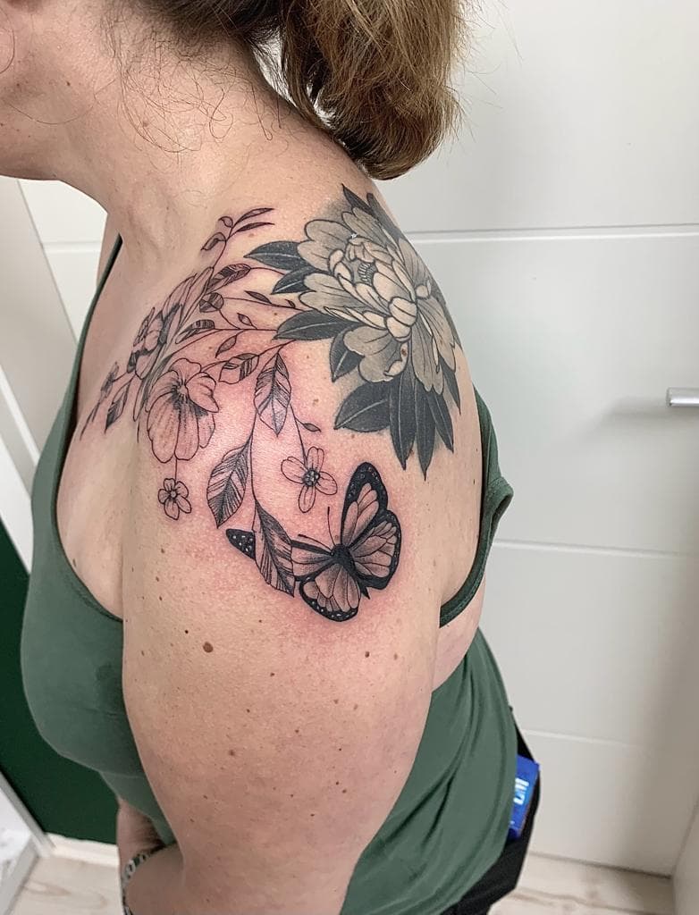 Zwart-wit tattoo van bloemen, bladeren en een vlinder op de schouder.