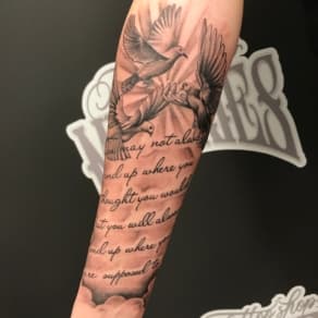 Zwart-wit tattoo van duiven, wolken en tekst op de onderarm.
