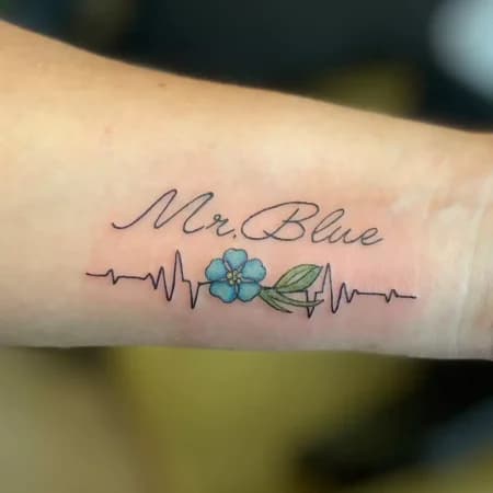 Minimalistische tattoo met 'Mr. Blue' tekst, hartslaglijn en blauw bloemetje.