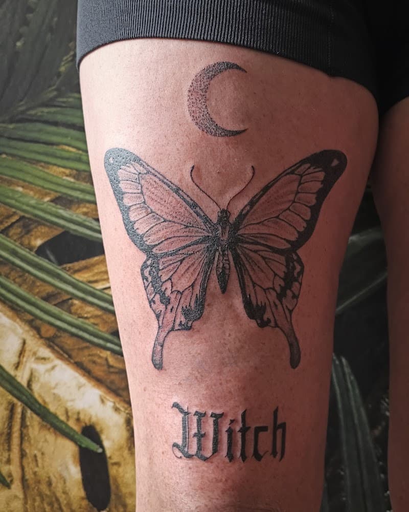 Zwart-wit tattoo van een vlinder, halve maan en 'Witch' op het bovenbeen.